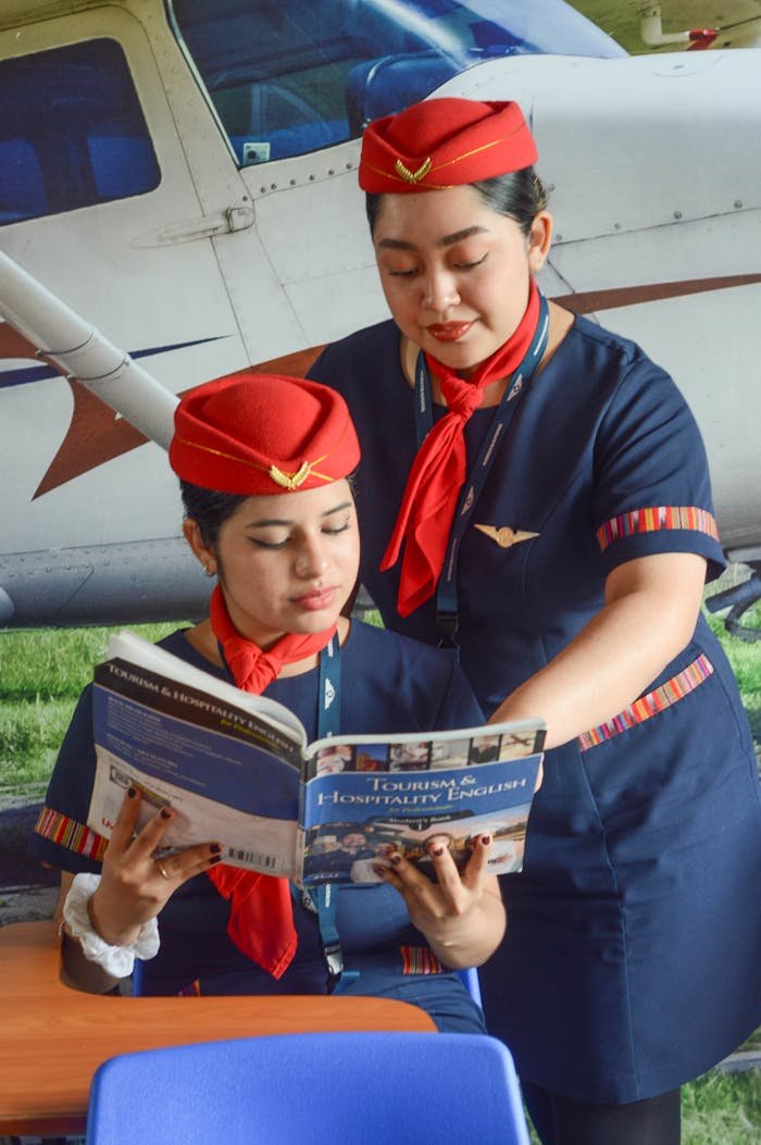 Two flight attendants in uniform studying hospitality English next to an airplane outdoors.