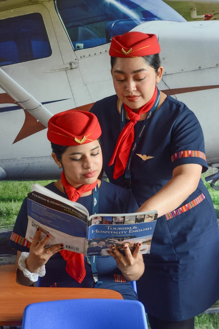 Two flight attendants in uniform reading aviation-related books outdoors near a plane.