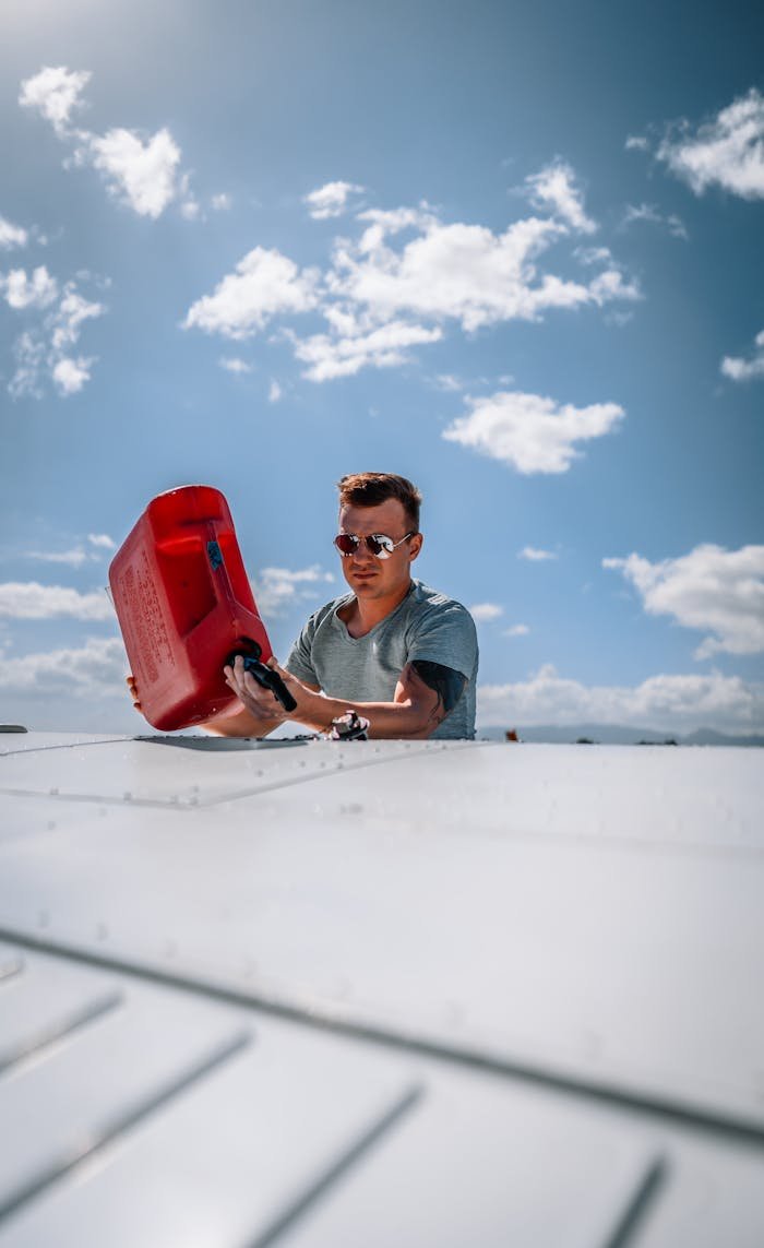 A man pours fuel into an airplane's fuel tank outdoors on a sunny day, wearing sunglasses for protection.
